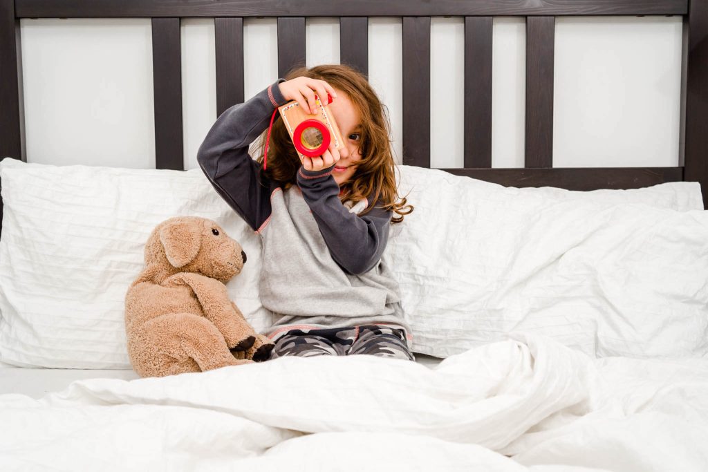 A young girl sits on a bed with her teddy modelling some ethnically produced pajamas. She is holding a wooden camera and pretending to take a photo just like Leicester commercial photographer Chris Bunce